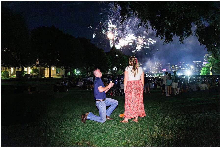A man proposes on one knee during Boston’s 4th of July fireworks, surrounded by a festive crowd—perfect for marriage proposal ideas Boston.