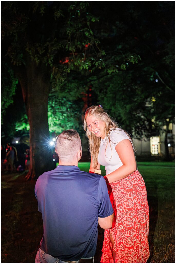 A close-up of a joyful proposal moment at night, with the woman smiling and the man on one knee holding a ring box.
