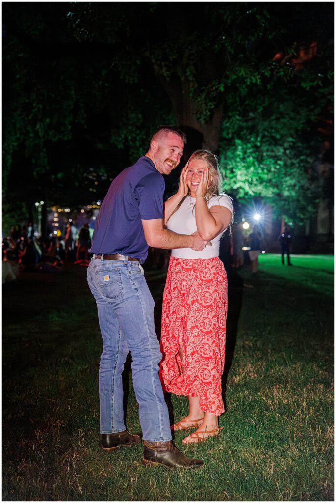 A couple laughs together on a grassy lawn at night after a proposal, the woman covering her face in joyful surprise.