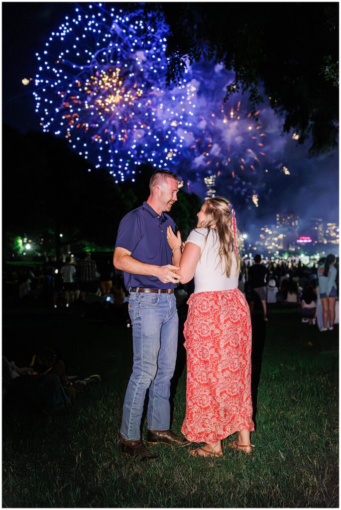 A happy couple holds hands under a sky filled with fireworks during a proposal, capturing one of the most memorable marriage proposal ideas Boston.