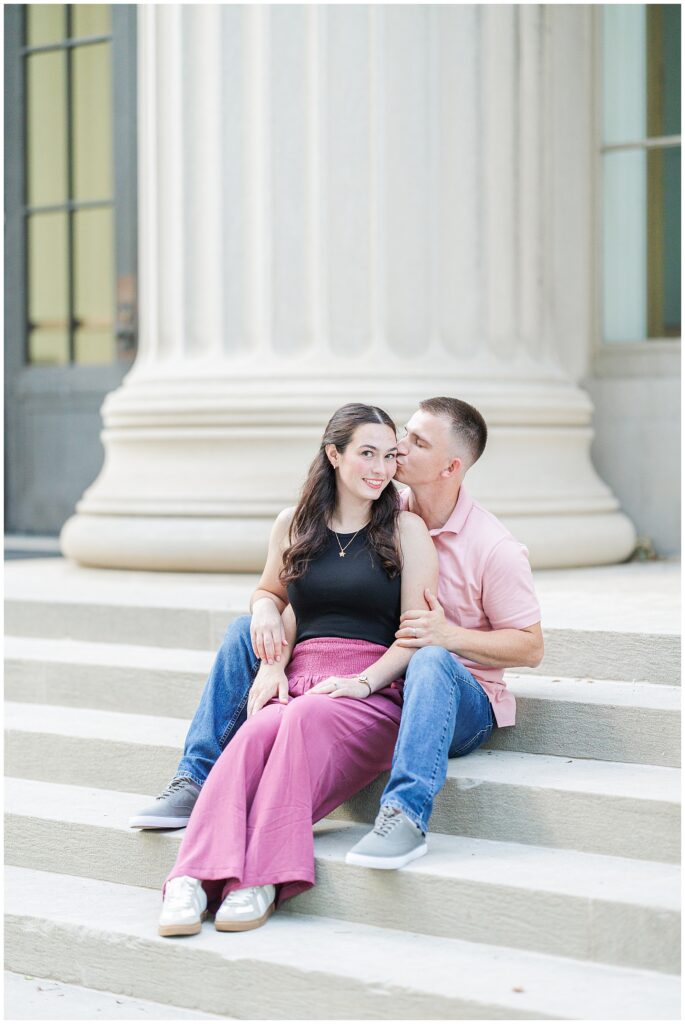A couple sits on stone steps in front of a neoclassical building with large columns. The man kisses the woman’s cheek as she smiles at the camera. Captured by a Cambridge photographer at MIT.