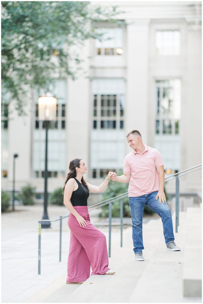 A couple stands on outdoor steps, holding hands and looking at each other playfully. The background shows a building with large windows and street lamps.
