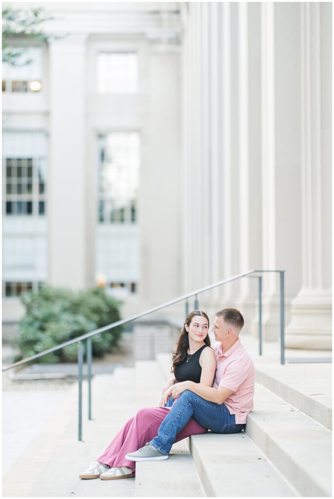 A couple sits on steps in front of large columns, leaning into each other with soft smiles. The woman looks at the man while he gazes forward.