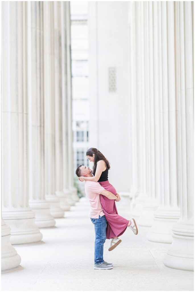 A man lifts a woman into the air between two rows of white columns at MIT, both smiling and making eye contact. Photo by a Cambridge photographer.