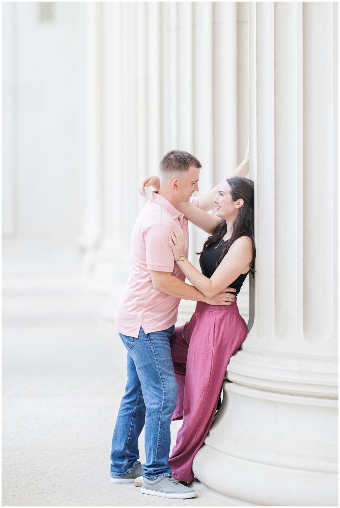 A couple embraces against a column. The woman leans against it with her arms around the man’s shoulders, and both are smiling.