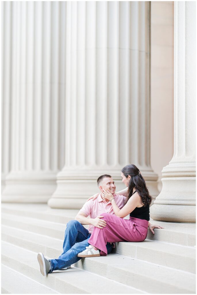 A couple sits on steps in front of classical columns. The woman gently holds the man’s face while he looks up at her, smiling.