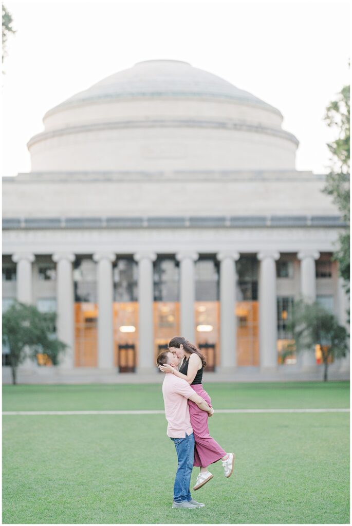 In front of MIT’s Great Dome, a man lifts a woman in a hug as they kiss, framed by symmetrical columns and evening light. Captured by a Cambridge photographer.
