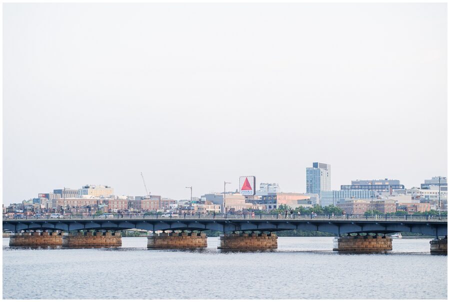A wide view of the Boston skyline at dusk, with the iconic Citgo sign and a long bridge crossing the Charles River. Captured by a Cambridge photographer from Memorial Drive.