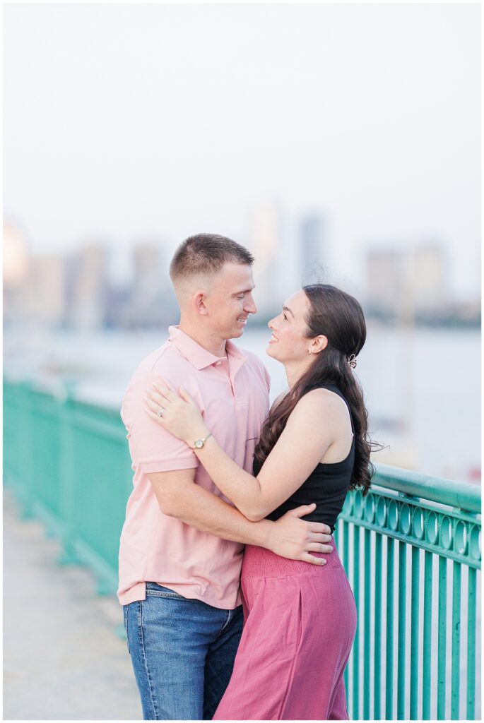 A couple stands close together on a riverside path, smiling and looking into each other’s eyes, with blurred city buildings in the background.