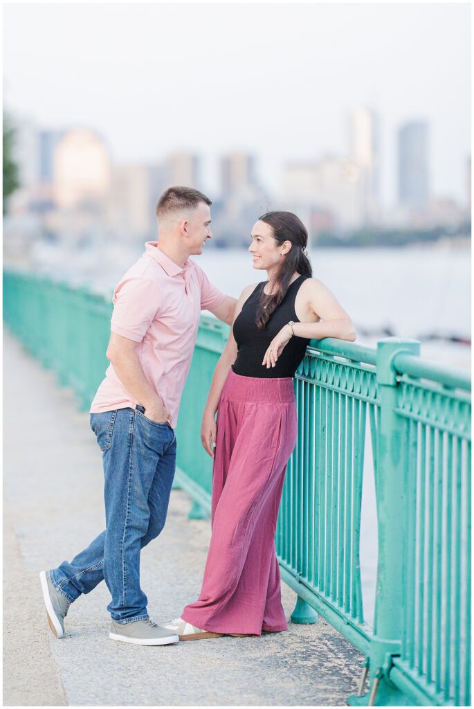 A couple leans casually on a green railing near the Charles River, smiling at each other with the city skyline in the background. Captured by a Cambridge photographer.