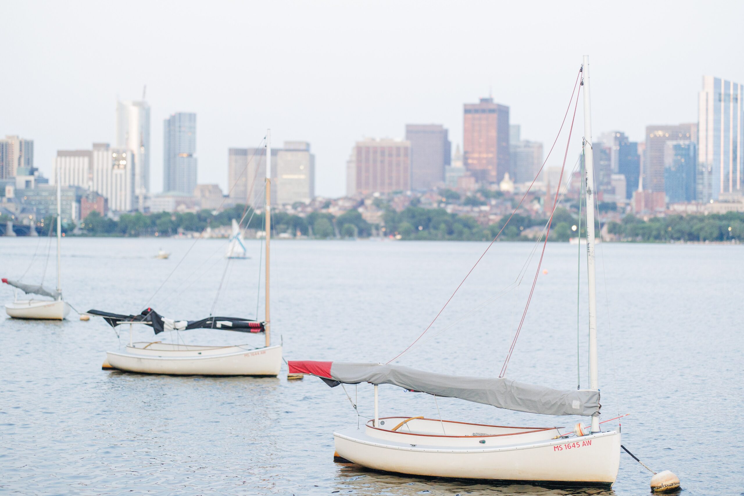 A close-up of a sailboat moored on the Charles River at dusk, with the Boston skyline in the background. Photo by a Cambridge photographer.