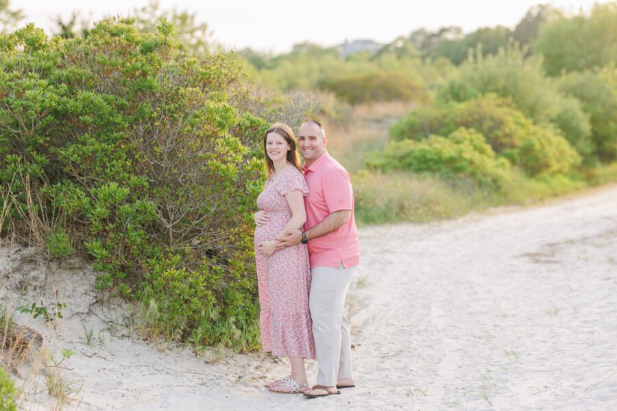 A couple poses on a beach path at Wingaersheek Beach, with the man embracing the pregnant woman from behind as they smile.