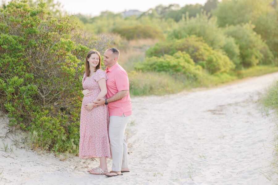 A pregnant woman in a floral dress stands on a beach path at Wingaersheek Beach, cradling her belly and smiling.