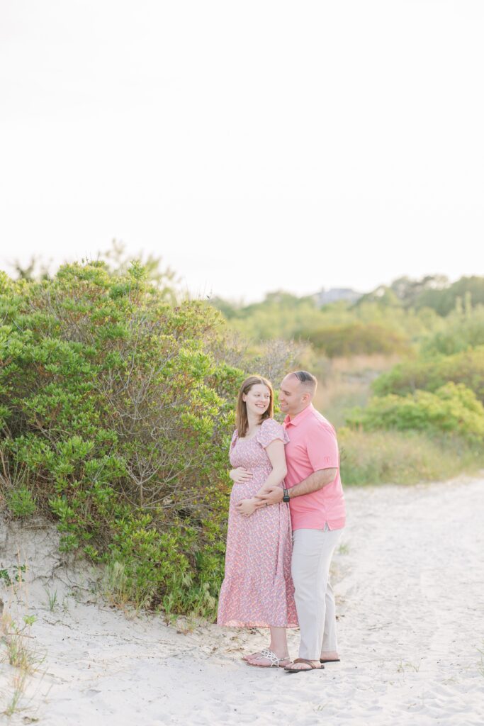 A pregnant woman and her partner walk together along a beach path at Wingaersheek Beach, holding hands and smiling.