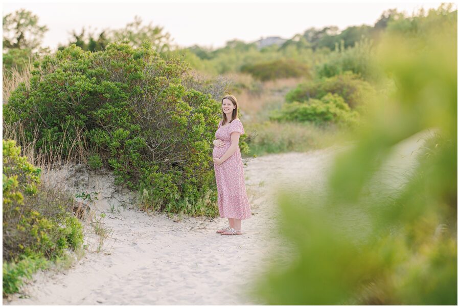Close-up of a pregnant woman’s hands on her belly in a pink floral dress at Wingaersheek Beach.