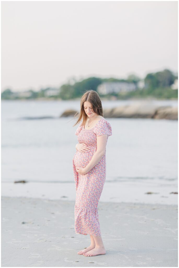A smiling pregnant woman stands on the beach at Wingaersheek, holding her belly in a floral dress.