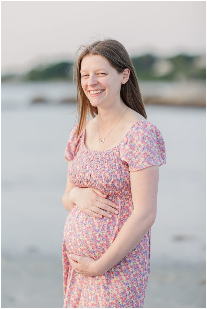 Pregnant woman smiling and holding her belly during a sunset session at Wingaersheek Beach.