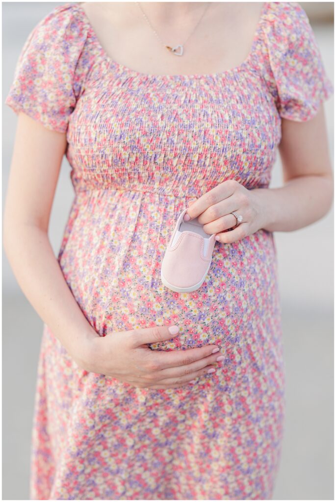 Close-up of a pregnant woman holding a tiny pink baby shoe over her belly in a floral dress.