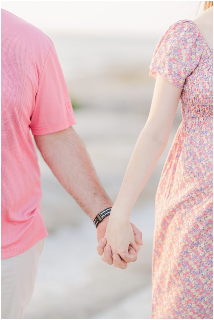 Close-up of a couple holding hands at Wingaersheek Beach, with the woman’s floral dress and man’s pink shirt visible.