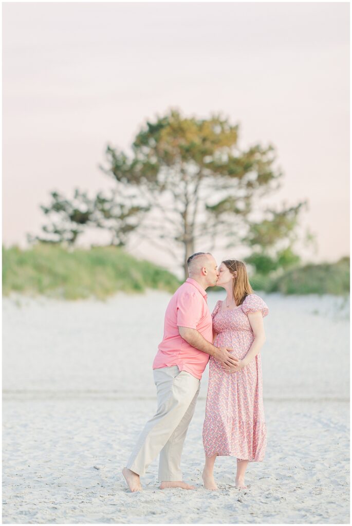 Expectant couple kissing on the beach with a tree in the background at Wingaersheek Beach.