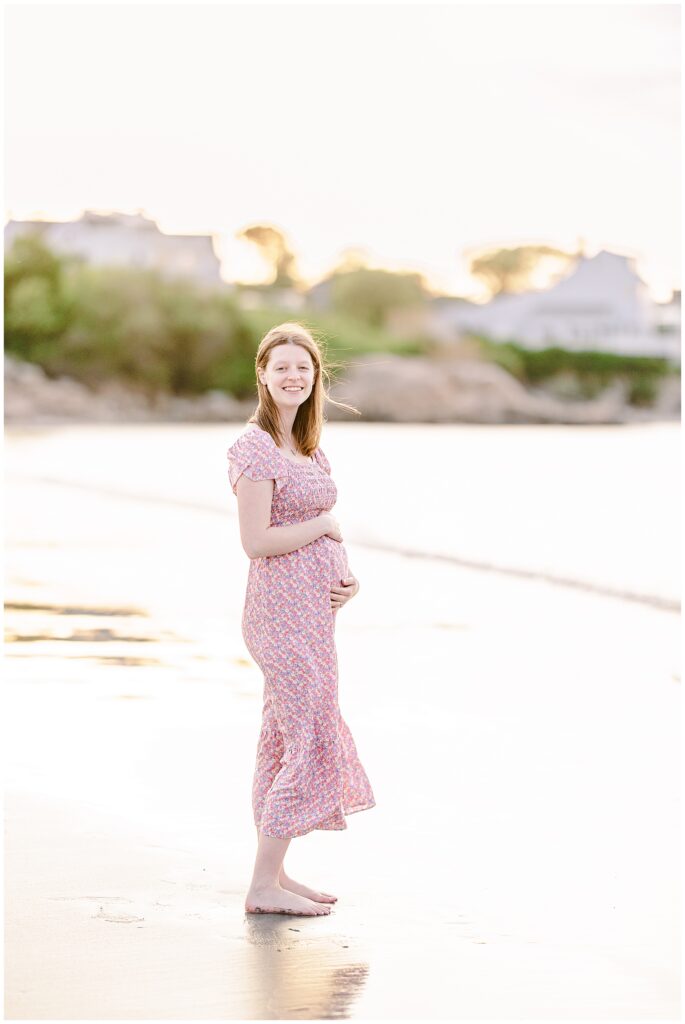 Pregnant woman smiling while standing barefoot on the beach at Wingaersheek, holding her belly.