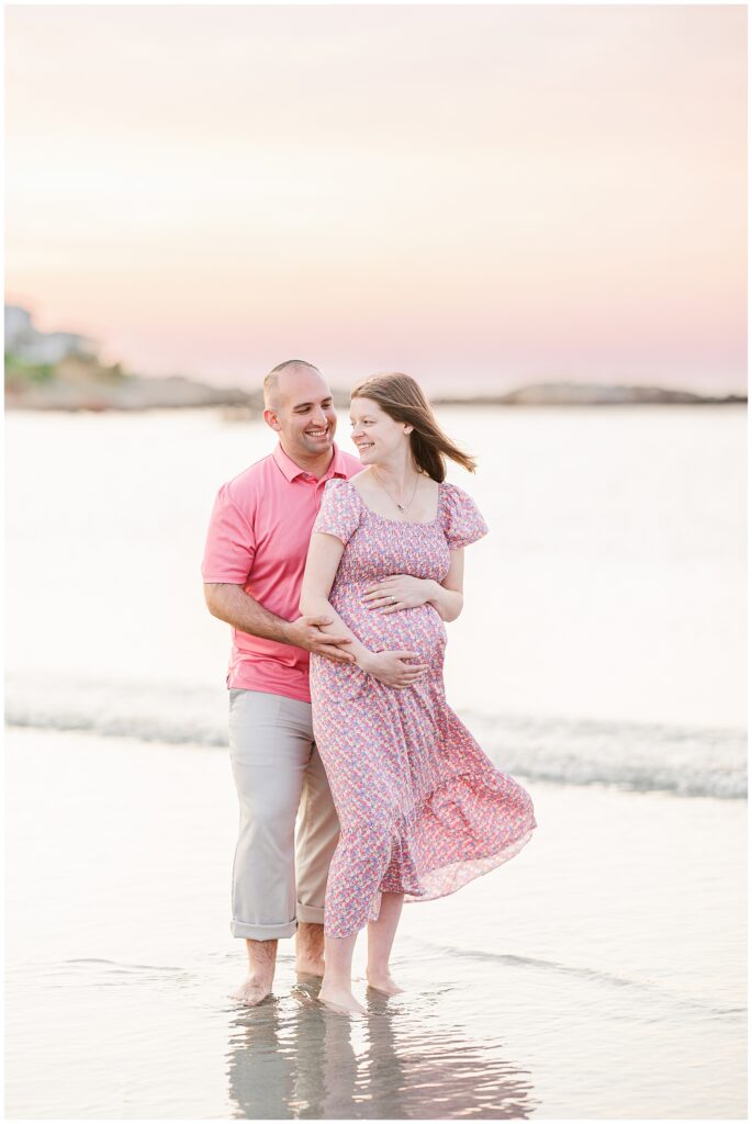 Expectant couple standing in the water at Wingaersheek Beach, smiling together at sunset.