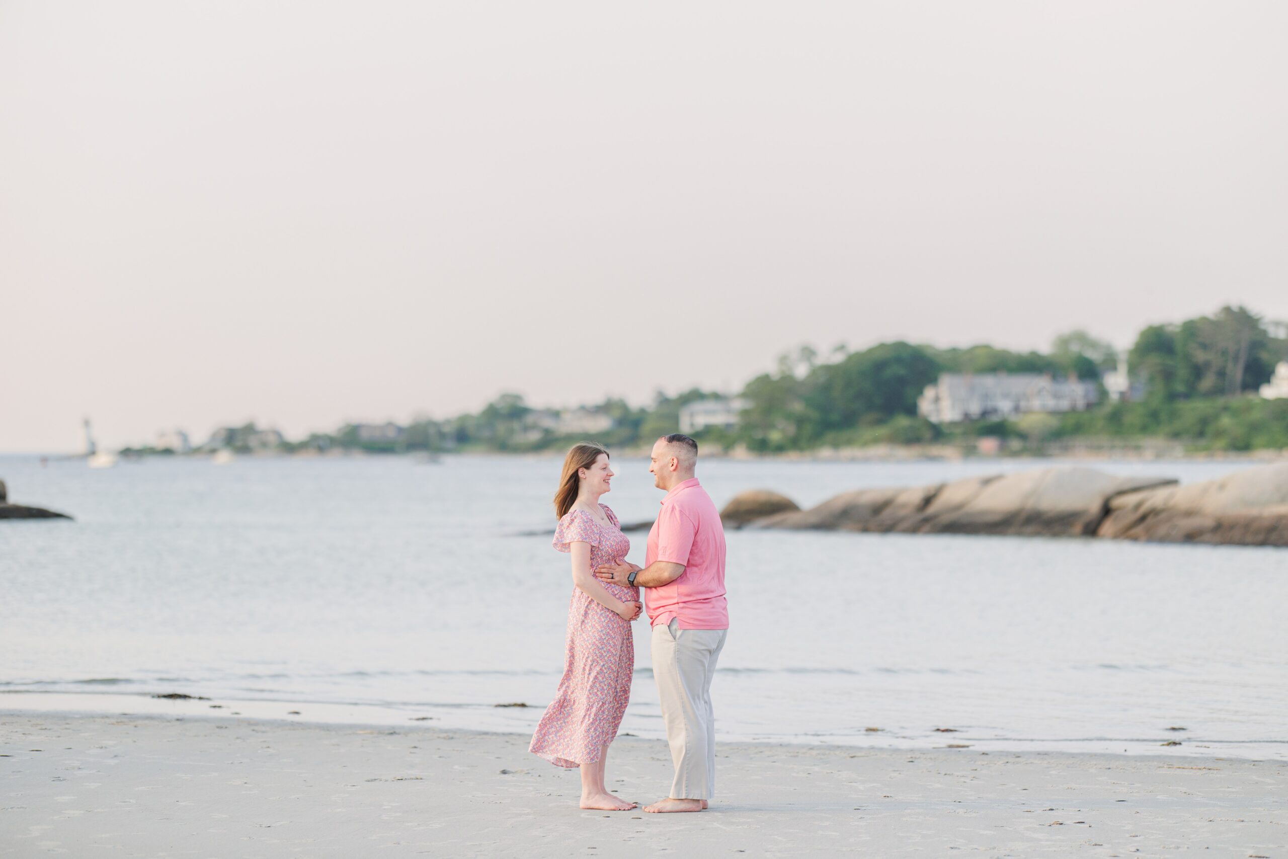 Maternity couple facing each other and smiling on the sand at Wingaersheek Beach in Gloucester.