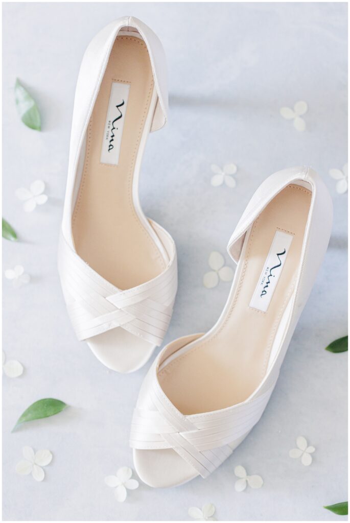 Ivory satin peep-toe bridal shoes styled flatlay with greenery and delicate white floral accents. A romantic bridal detail photographed by a Cape Cod wedding photographer.