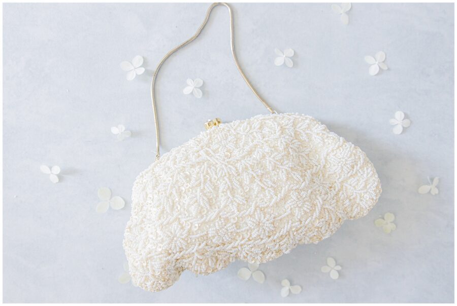 Beaded ivory bridal clutch with floral patterning styled on a light background with petals. A timeless accessory image captured by a Cape Cod wedding photographer.