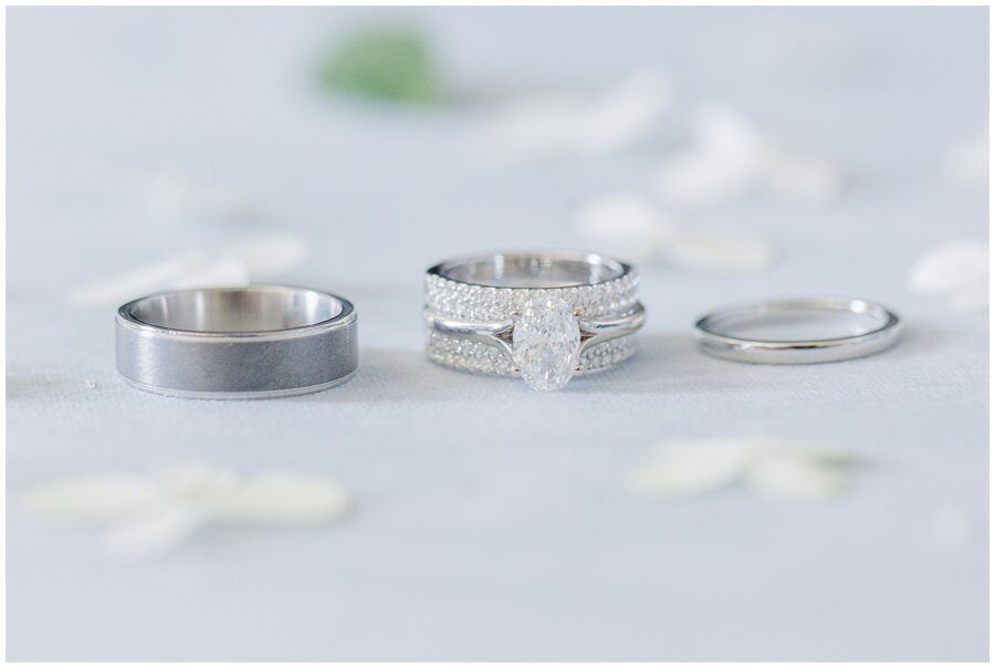 Trio of wedding rings displayed on soft linen with scattered white petals for a modern flatlay. Elegant bridal details photographed on Cape Cod.