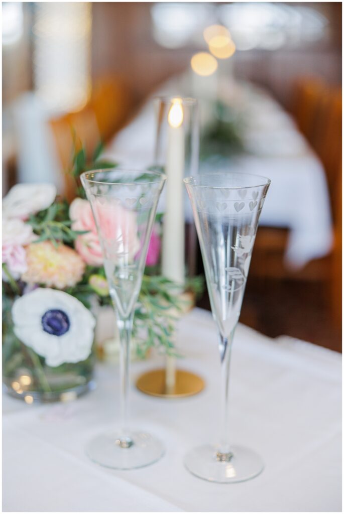 Etched champagne flutes with heart designs displayed on a wedding reception table with floral décor. A joyful wedding detail image captured by a Cape Cod wedding photographer.