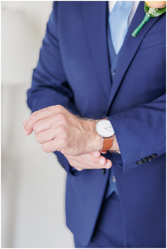Close-up of groom’s hands adjusting his wristwatch while wearing a navy suit and boutonniere. A timeless groom detail image on Cape Cod.