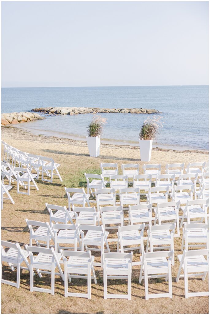 Ceremony setup with rows of white chairs facing the ocean, framed by tall floral arrangements. A seaside Cape Cod wedding ceremony space captured by a Cape Cod wedding photographer.