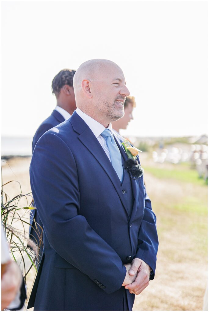 Groom smiling at the altar in a navy suit with a light blue tie, waiting for his bride by the ocean. A candid Cape Cod wedding ceremony moment captured by a Cape Cod wedding photographer.