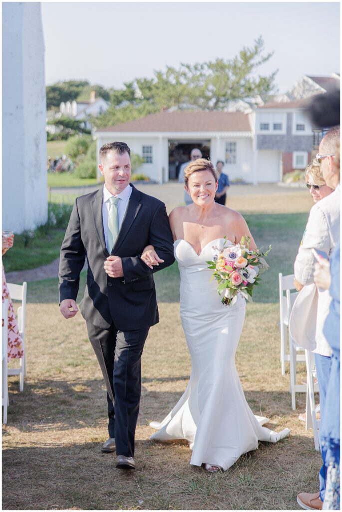 Bride walking down the aisle in a strapless gown, holding a bouquet, escorted by a family member. A joyful seaside Cape Cod wedding ceremony.