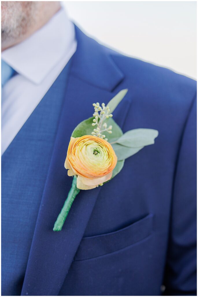 Close-up of groom’s boutonniere, a yellow ranunculus accented with greenery, pinned to a navy suit. A refined wedding detail image captured by a Cape Cod wedding photographer.