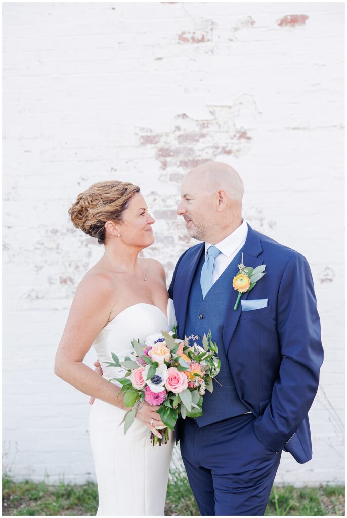Bride and groom standing together against a rustic white brick wall, smiling into each other’s eyes. A timeless Cape Cod wedding portrait.