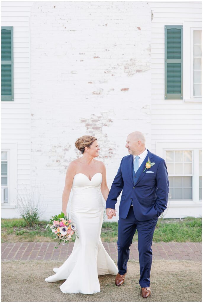 Bride and groom walking hand-in-hand, smiling at one another, in front of a historic white building. A romantic wedding day moment on Cape Cod.