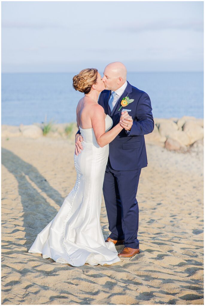Bride and groom kissing on the beach, framed by the ocean and golden evening light. A romantic waterfront wedding image captured by a Cape Cod wedding photographer.