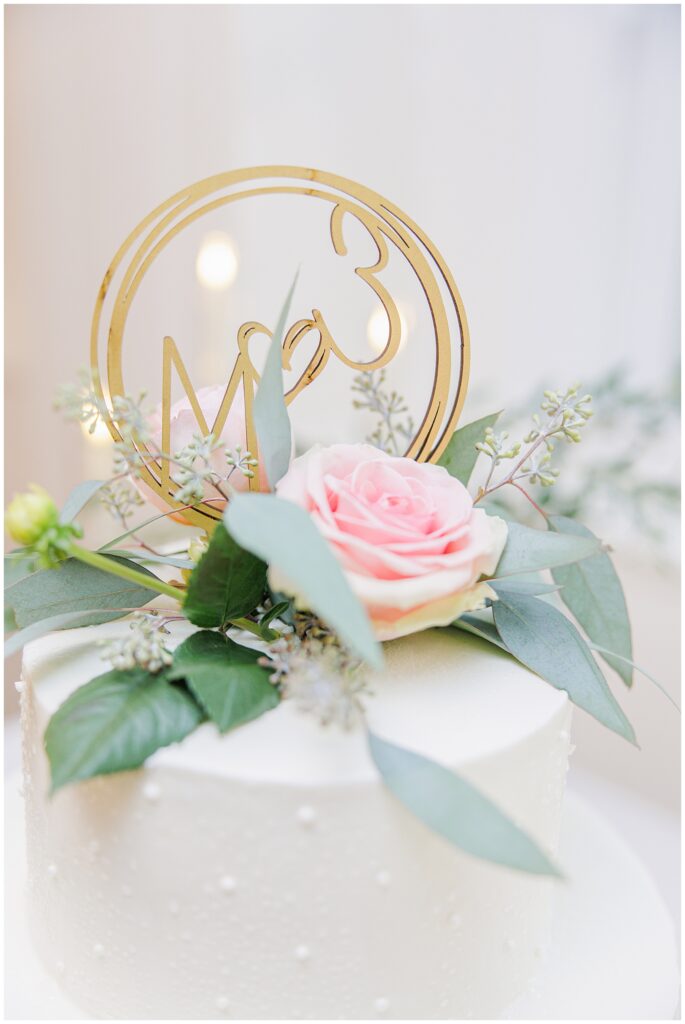 Close-up of wedding cake topper with blush rose, greenery, and gold “Mr & Mrs” design. Elegant reception detail photographed on Cape Cod.