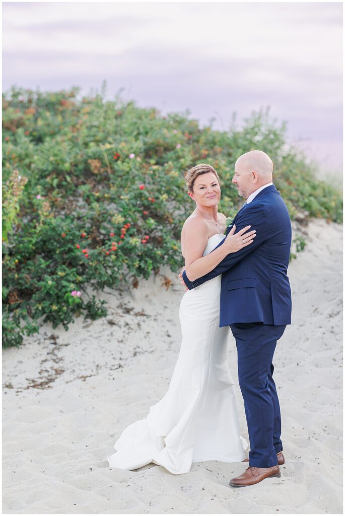 Bride and groom embracing on the sand dunes with greenery in the background. A romantic Cape Cod wedding portrait at sunset.