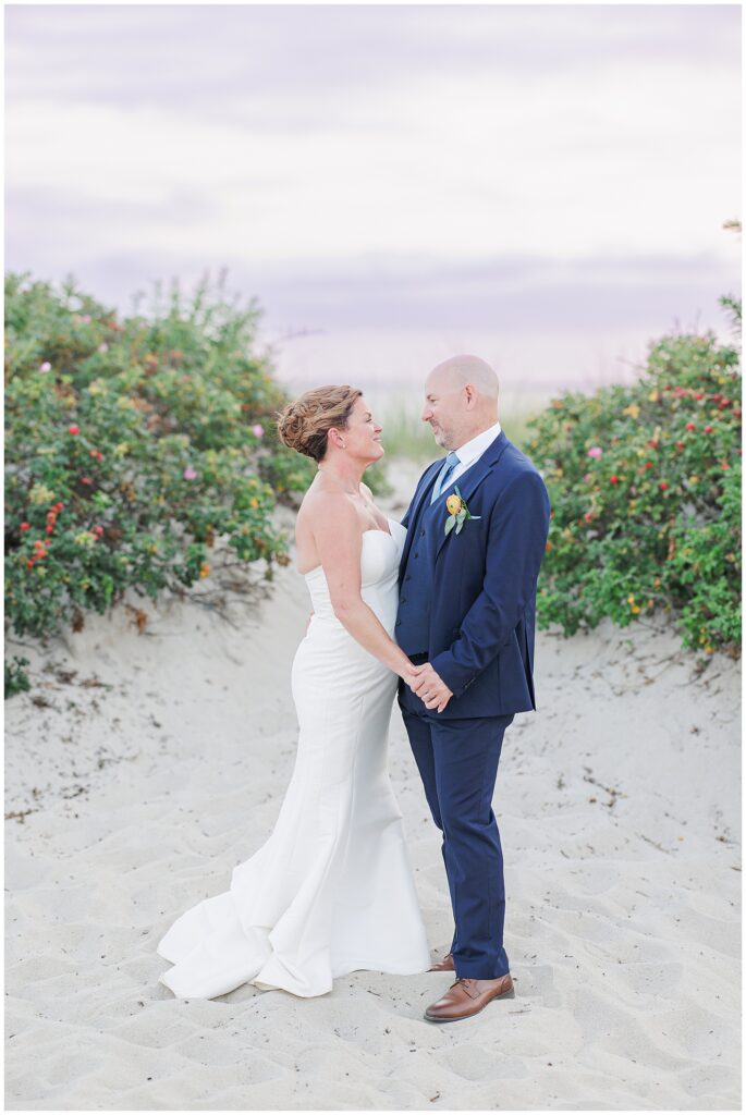 Bride and groom holding hands and smiling at each other on the sand dunes. A timeless Cape Cod wedding portrait captured by a Cape Cod wedding photographer.