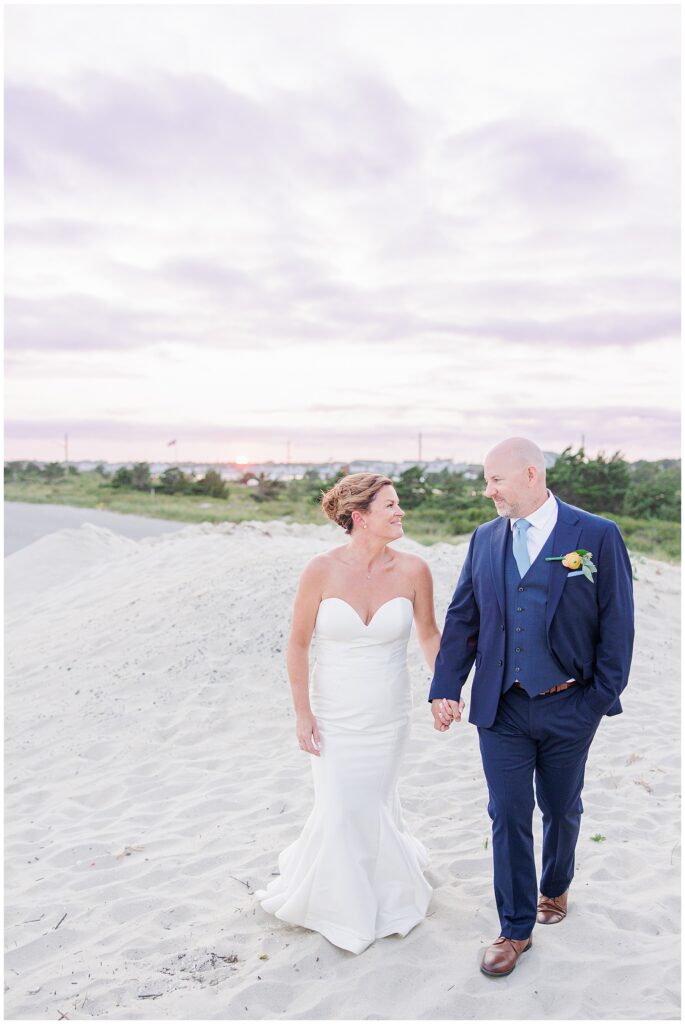 Bride and groom walking hand in hand on the beach at sunset, smiling toward each other. A romantic seaside Cape Cod wedding image.