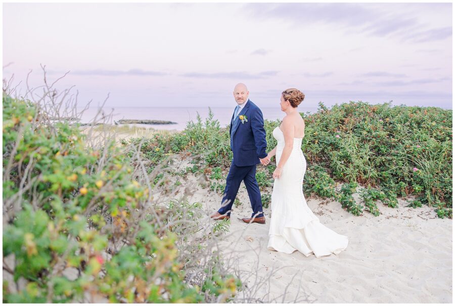 Bride and groom holding hands, walking along the sand dunes with greenery in the background. A candid Cape Cod wedding photographer capture.