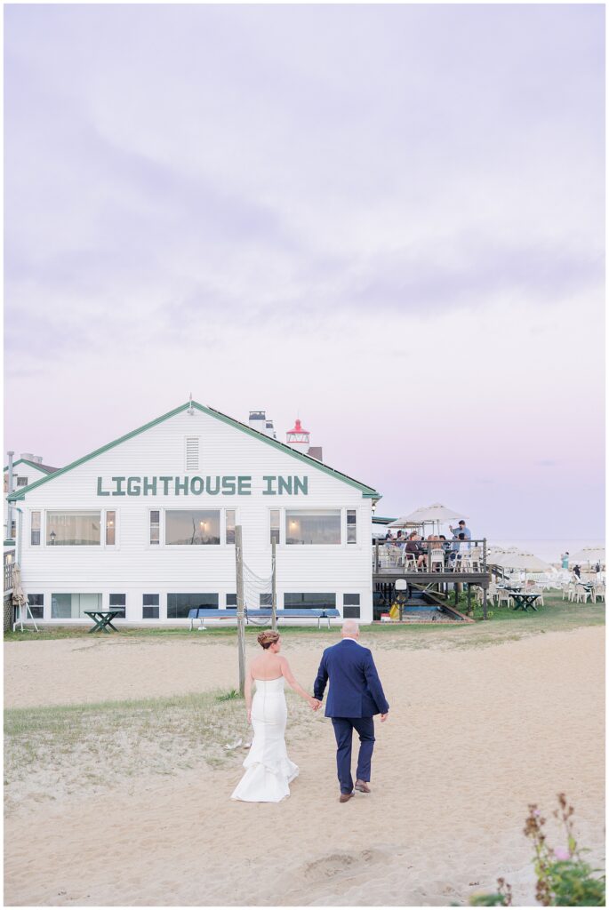 Bride and groom walking hand in hand toward a white seaside inn at sunset. A relaxed Cape Cod wedding moment captured by a Cape Cod wedding photographer.