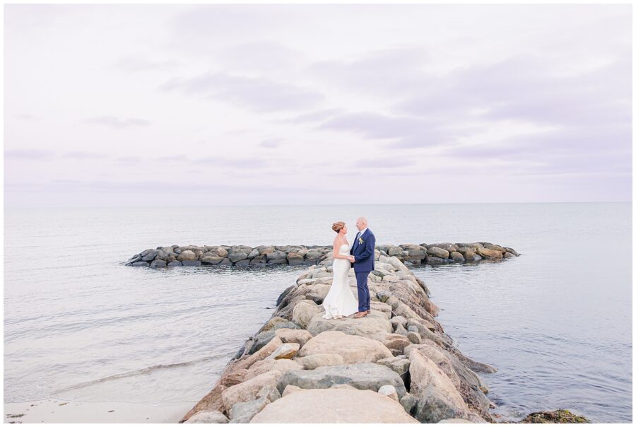 Bride and groom standing together on a rock jetty surrounded by ocean waves. A cinematic Cape Cod waterfront wedding portrait.