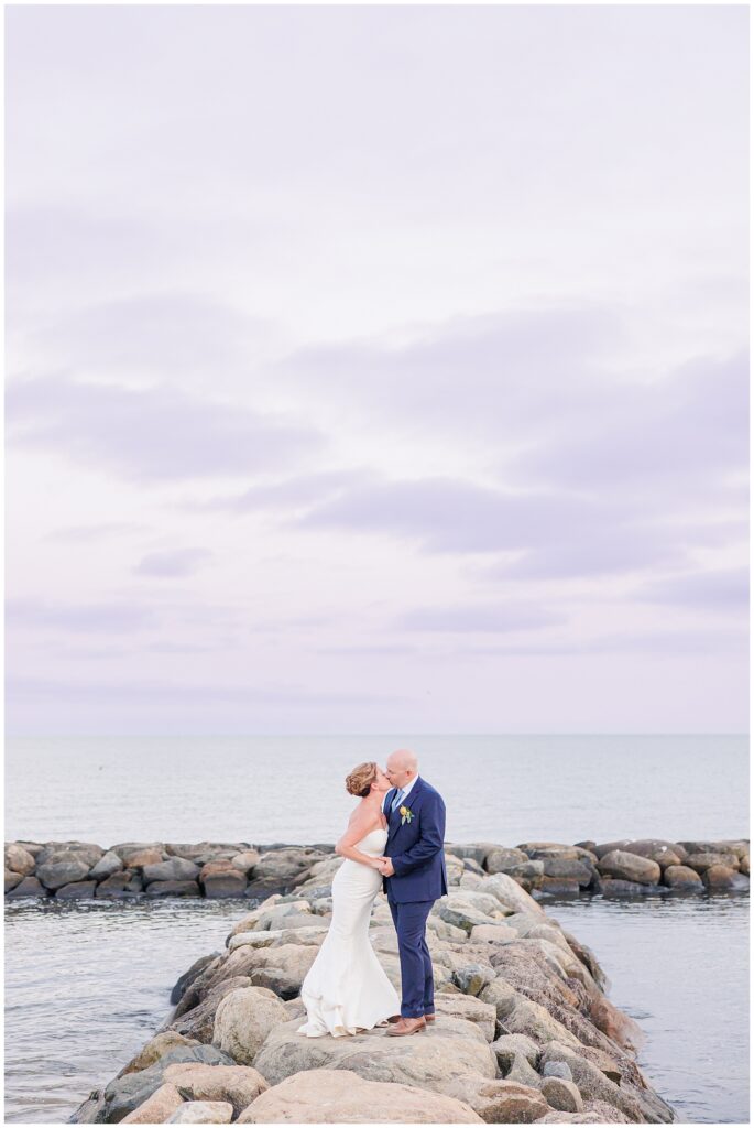 Bride and groom kissing on a rocky jetty with the ocean stretching behind them. A romantic seaside Cape Cod wedding image captured by a Cape Cod wedding photographer.
