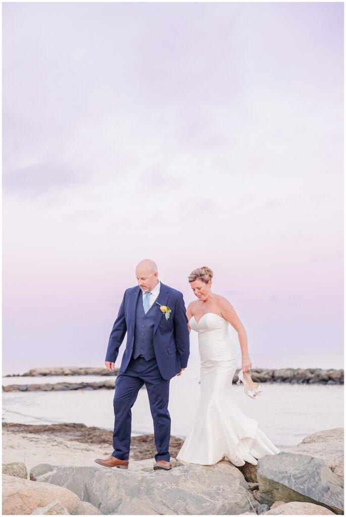 Bride and groom walk carefully across rocks at the Cape Cod shoreline during sunset, with the sky glowing in shades of purple and pink. The bride holds her shoes in one hand while the groom leads her forward, representing the portfolio of a Cape Cod wedding photographer.
