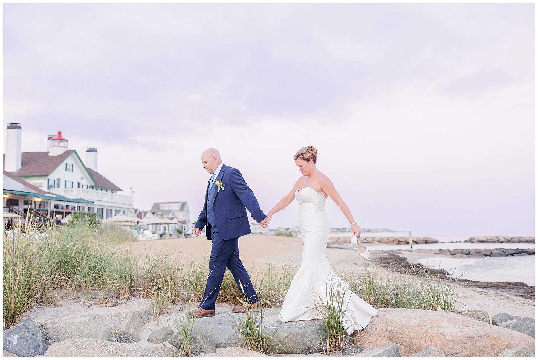 Bride and groom hold hands while walking along the Cape Cod beach near dune grasses, with the classic New England inn and lighthouse in the background under a pastel sunset sky, representing the portfolio of a Cape Cod wedding photographer.