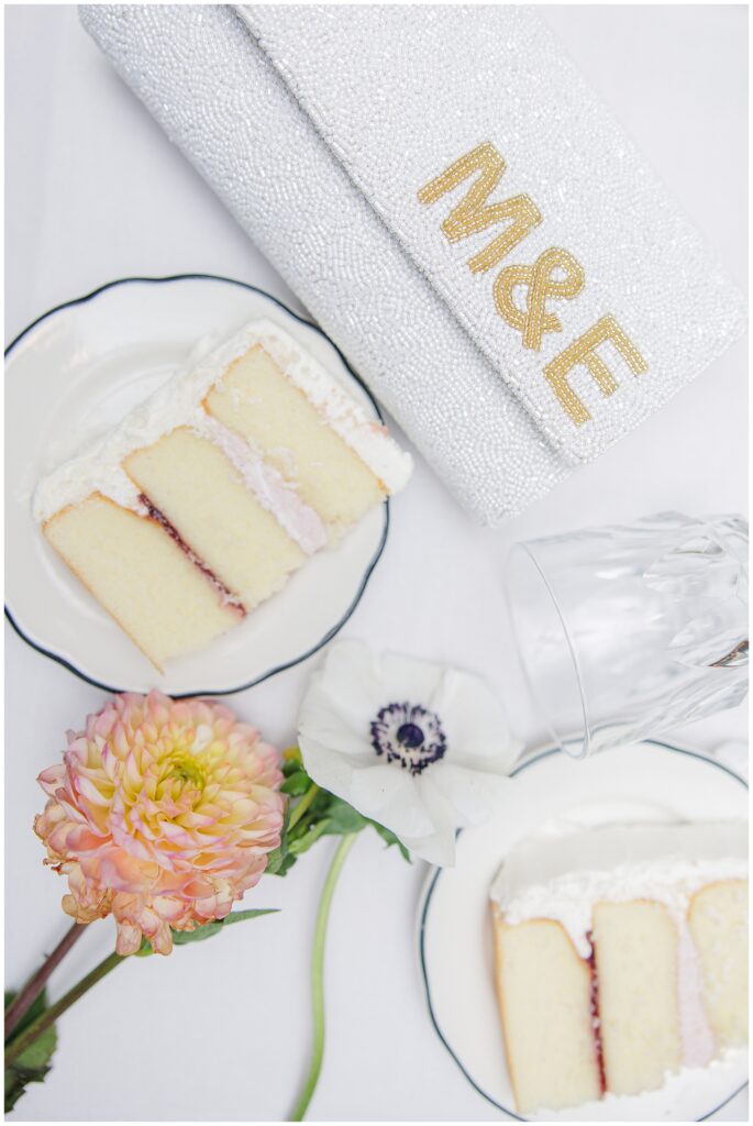 Close-up of two slices of white wedding cake with berry filling, styled alongside a white beaded clutch embroidered with “M&E” and fresh floral accents, representing the portfolio of a Cape Cod wedding photographer.